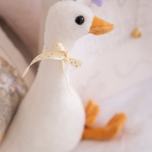 May include: A white plush goose toy with a yellow beak and a gold polka dot ribbon around its neck. The goose is sitting on a white surface.