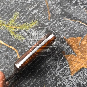 May include: A close-up of a brown and silver knife with a wooden handle. The knife is on a black and white background with a pattern of leaves and the text "Hinterland Knives" repeated.