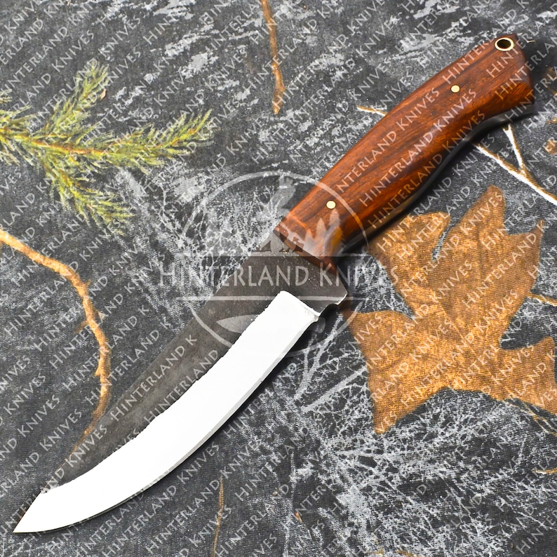 May include: A white-bladed hunting knife with a brown wooden handle. The knife has a curved blade and a brass pin on the handle. The knife is on a camouflage background.