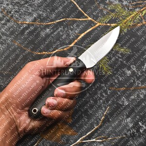 May include: A black handled knife with a plain, silver blade. The knife is held in a hand against a camouflage background.