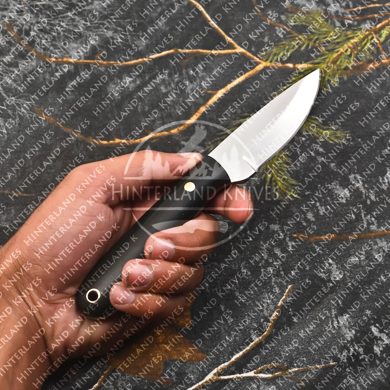 May include: A black handled knife with a plain, silver blade. The knife is held in a hand against a camouflage background.