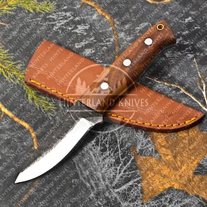 Puede incluir: Un cuchillo pequeño con mango marrón, hoja curva y funda de cuero. El cuchillo tiene un remache de latón en el mango y la funda tiene las palabras "Hinterland Knives" grabadas.
