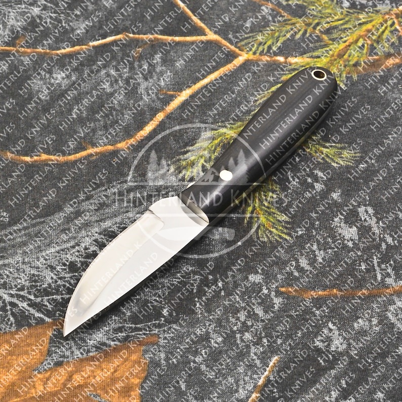 May include: A small, silver knife with a black wooden handle. The knife has a pointed blade and a small, round rivet on the handle. The knife is lying on a camouflage fabric.