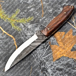 May include: A hunting knife with a brown wooden handle and a curved, partially-polished blade. The knife is lying on a camouflage-patterned fabric.