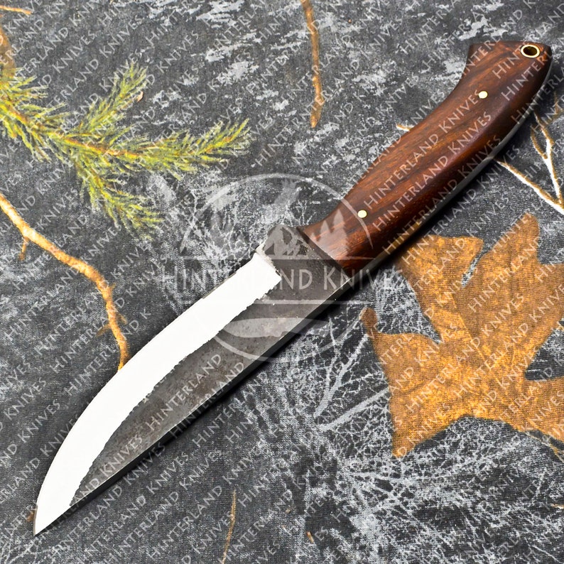 May include: A hunting knife with a brown wooden handle and a curved, partially-polished blade. The knife is lying on a camouflage-patterned fabric.