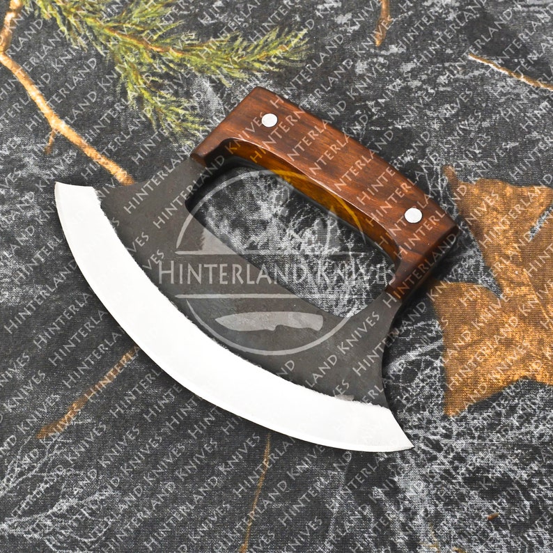 May include: A curved, stainless steel knife with a wooden handle. The knife has a white blade and a brown handle with a light brown accent. The knife is on a camouflage fabric background. The text "Hinterland Knives" is printed on the blade.