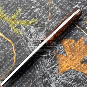 May include: A close-up of a knife with a wooden handle and a silver blade. The handle has a white stripe running down the center. The blade is sharp and pointed. The knife is on a black and white camouflage background. The text "Hinterland Knives" is printed on the handle.