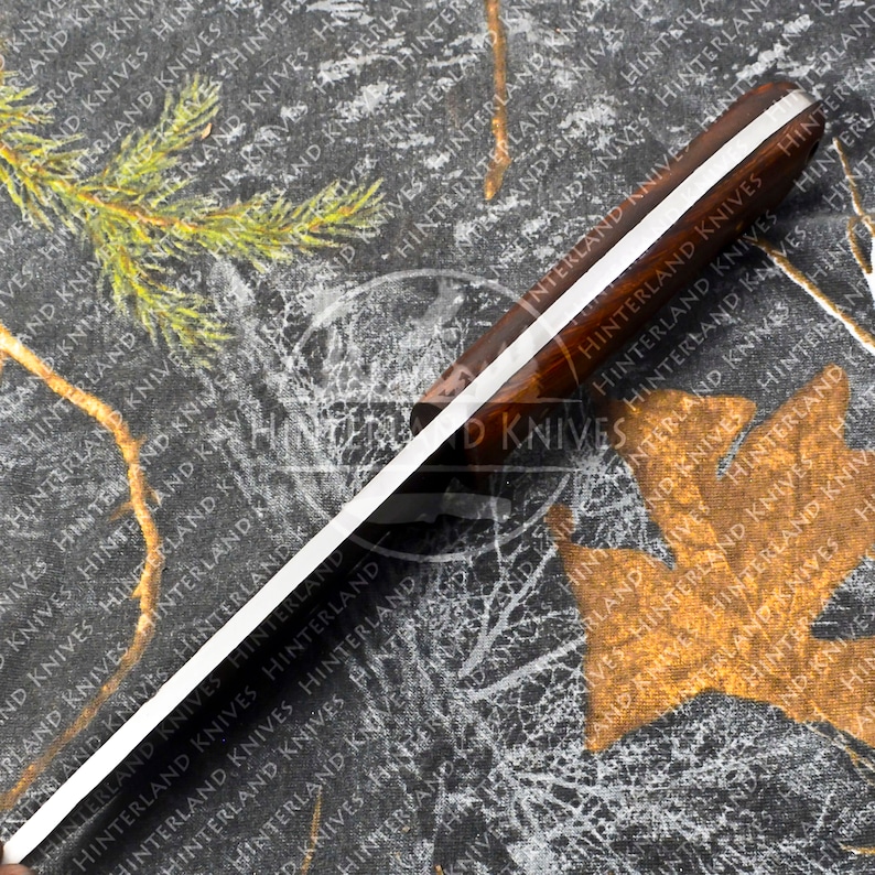 May include: A close-up of a knife with a wooden handle and a silver blade. The handle has a white stripe running down the center. The blade is sharp and pointed. The knife is on a black and white camouflage background. The text "Hinterland Knives" is printed on the handle.