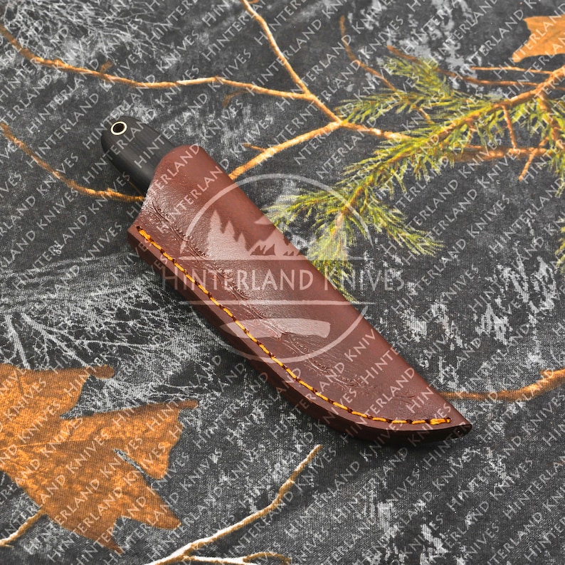 May include: A brown leather knife sheath with yellow stitching. The sheath has the text "Hinterland Knives" embossed on the front.
