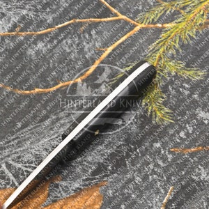 May include: A black and white hunting knife with a black handle and a white stripe running down the center of the blade. The knife is lying on a camouflage fabric background.