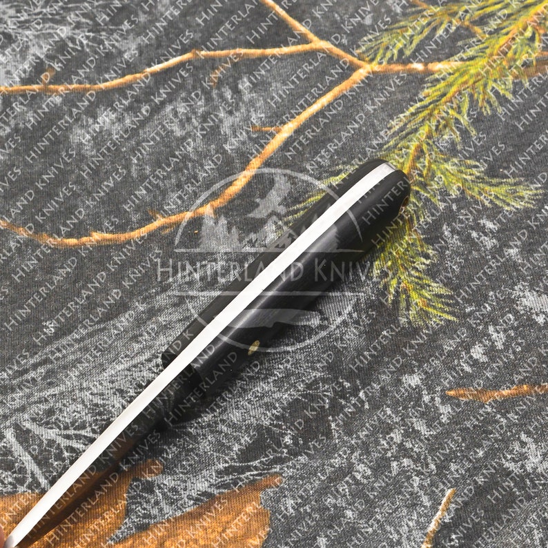 May include: A black and white hunting knife with a black handle and a white stripe running down the center of the blade. The knife is lying on a camouflage fabric background.