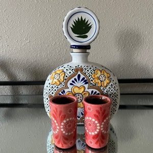 May include: A white ceramic tequila bottle with a green agave plant design on the stopper. Two small pink ceramic shot glasses with white floral designs are in front of the bottle.