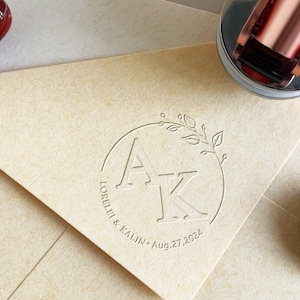 Puede incluir: Sobre de invitación de boda color crema con un diseño de monograma en relieve con las letras "AK" dentro de una corona circular. También se incluye la fecha "27 de agosto de 2024". Se ve un sello de cera de oro rosa.