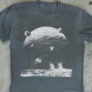 以下が含まれることがあります： ヴィンテージ加工が施されたグレーのTシャツに、白いクモの巣、クモ、惑星のグラフィックプリントが施されています。シャツの上部には白い文字で「DISTRESSED VINTAGE」とプリントされています。