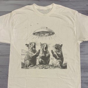 May include: A white t-shirt with a graphic of three squirrels looking up at a flying saucer. The text "SAND" is in the upper right corner of the image.