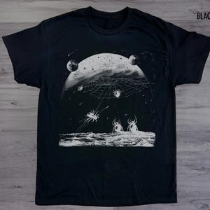 以下が含まれることがあります： 月のクモの巣とクモの白いグラフィックプリントが施された黒いTシャツ。グラフィックはヴィンテージスタイルです。