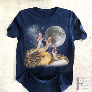 May include: Navy blue t-shirt featuring a graphic of three snakes with open mouths, set against a starry night sky and a full moon. The snakes are in shades of brown and yellow. The shirt has a crew neck.