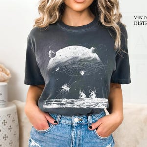 以下が含まれることがあります： ヴィンテージ加工が施されたダークグレーのTシャツ。白い月とクモの巣、クモが描かれたグラフィックデザインです。