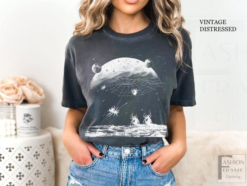 以下が含まれることがあります： ヴィンテージ加工が施されたダークグレーのTシャツ。白い月とクモの巣、クモが描かれたグラフィックデザインです。