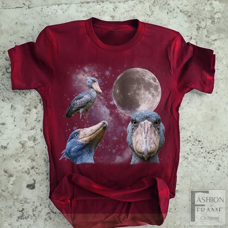 May include: A maroon t-shirt featuring a graphic design of a Shoebill stork with a full moon and a starry background. The design includes three different views of the bird. The shirt has a crew neck and short sleeves.