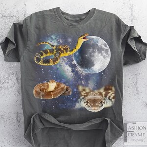 May include: Dark gray t-shirt featuring a vibrant graphic of snakes, a full moon, and a starry galaxy background. The design includes three different snake illustrations. The shirt has short sleeves and a crew neck.