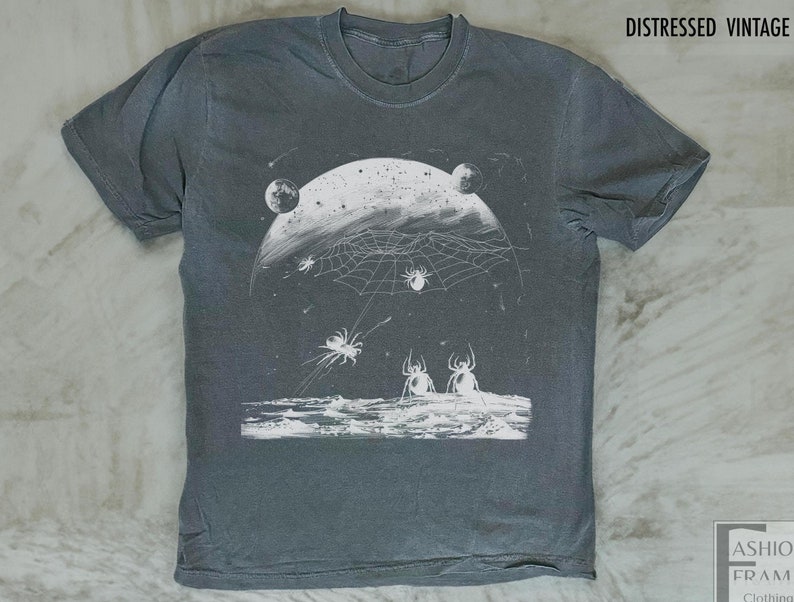 以下が含まれることがあります： ヴィンテージ加工が施されたグレーのTシャツに、白いクモの巣、クモ、惑星のグラフィックプリントが施されています。シャツの上部には白い文字で「DISTRESSED VINTAGE」とプリントされています。