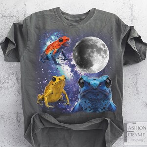May include: Dark gray t-shirt featuring a graphic of three colorful frogs against a space-themed background with a full moon. The frogs are orange, yellow, and blue. The shirt has a distressed look.