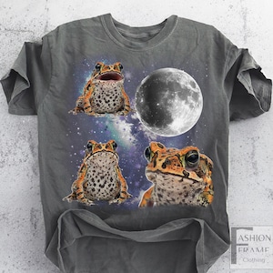 May include: Gray t-shirt featuring a graphic design of three toads and a full moon against a starry galaxy background. The toads have orange and brown markings. The shirt has a distressed look.