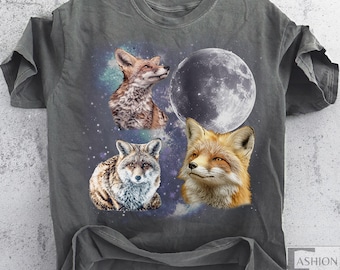 May include: A gray t-shirt featuring a design with three fox portraits and a full moon against a starry background. The foxes are depicted in shades of brown, orange, and gray. The shirt has a relaxed fit and short sleeves.