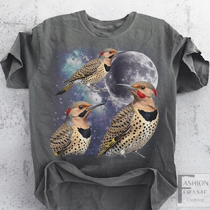 May include: Gray t-shirt featuring a vibrant illustration of three Northern Flicker birds with a full moon and a starry galaxy background. The birds have red markings, black bibs, and spotted plumage. The shirt has a relaxed fit.