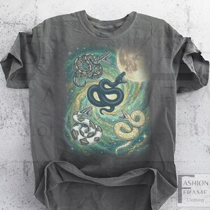 May include: A gray t-shirt featuring a colorful graphic of four snakes in a cosmic setting with a full moon. The snakes are in shades of black, white, and yellow, with the background in teal, yellow, and green.