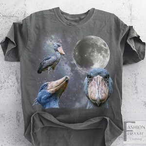 May include: Dark gray t-shirt featuring a graphic design of a Shoebill stork, a full moon, and a starry background. The shirt has a crew neck and short sleeves. The text "FASHION FRAME CLOTHING" is visible on the bottom right.