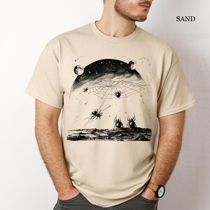 以下が含まれることがあります： 砂色のTシャツに、星が輝く夜空に蜘蛛の巣と蜘蛛が描かれた白黒のグラフィックプリントが施されています。画像の右上に「SAND」という文字が印刷されています。