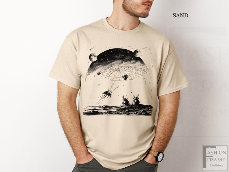 以下が含まれることがあります： 砂色のTシャツに、星が輝く夜空に蜘蛛の巣と蜘蛛が描かれた白黒のグラフィックプリントが施されています。画像の右上に「SAND」という文字が印刷されています。