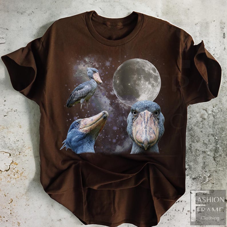 May include: Brown t-shirt featuring a graphic design of three shoebill storks, a full moon, and a starry background. The shoebills are depicted in various poses, with detailed feather textures and distinctive beaks. The shirt has a crew neck and short sleeves.