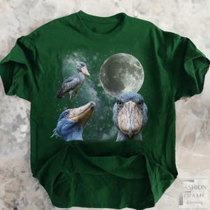 May include: A dark green t-shirt featuring a design with three Shoebill Stork illustrations, a full moon, and a starry background. The Shoebills are in various poses, with detailed depictions of their beaks and feathers. The shirt has a Fashion Frame Clothing logo.