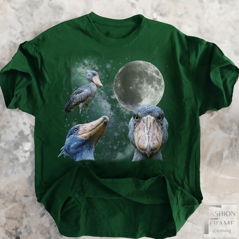 May include: A dark green t-shirt featuring a design with three Shoebill Stork illustrations, a full moon, and a starry background. The Shoebills are in various poses, with detailed depictions of their beaks and feathers. The shirt has a Fashion Frame Clothing logo.