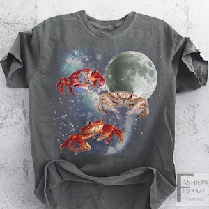 May include: Gray t-shirt featuring a celestial design with three crabs, a full moon, and a starry background. The crabs are red and tan, with the moon in shades of gray. The shirt has a relaxed fit.