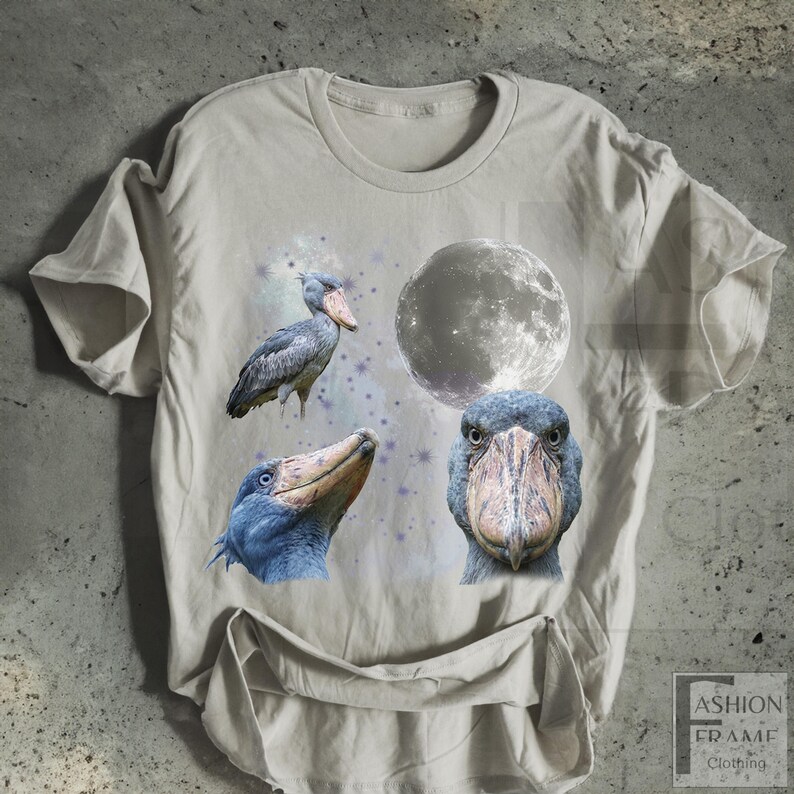 May include: A light beige t-shirt featuring a graphic design of a Shoebill stork, a full moon, and stars. The Shoebill stork is depicted in three different views. The shirt has a crew neck and short sleeves.
