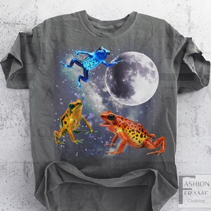 May include: Gray t-shirt featuring a vibrant graphic of three colorful frogs against a cosmic background with a full moon. One frog is blue, another is orange, and the third is yellow. The shirt has the text "FASHION FRAME CLOTHING" at the bottom.