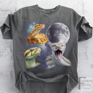 May include: Gray t-shirt featuring a graphic design of three snakes and a full moon against a starry background. The snakes are in shades of brown, green, and white. The shirt has a classic crew neck and short sleeves.