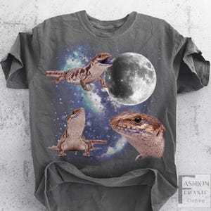 May include: Gray t-shirt featuring a graphic of three lizards with a moon and a starry galaxy background. The lizards are brown and tan, with one appearing to be biting the moon. The shirt has a distressed look.