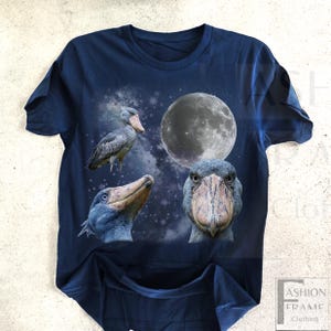 May include: Navy blue t-shirt featuring a graphic design of a Shoebill stork with a full moon and starry background. The design includes three different views of the bird. The text "FASHION FRAME CLOTHING" is visible in the lower right corner.
