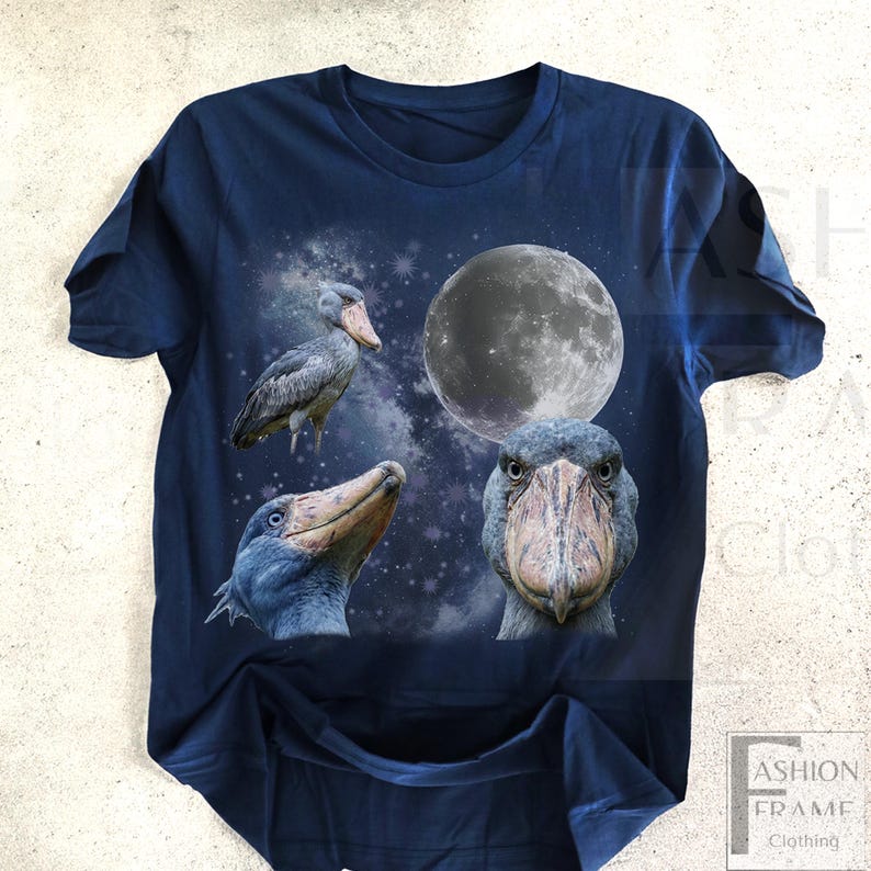 May include: Navy blue t-shirt featuring a graphic design of a Shoebill stork with a full moon and starry background. The design includes three different views of the bird. The text "FASHION FRAME CLOTHING" is visible in the lower right corner.