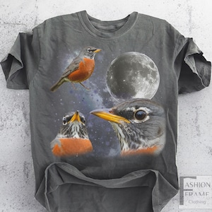 May include: Gray t-shirt featuring a graphic design of three robins and a full moon against a starry background. The robins have orange breasts and yellow beaks. The shirt has a distressed look.