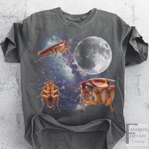 May include: Dark gray t-shirt featuring a graphic design of three cockroaches, a full moon, and a starry galaxy background. The shirt has a crew neck and short sleeves. The text "FASHION FRAME CLOTHING" is in the lower right corner.