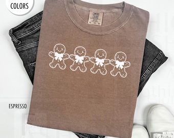 May include: A brown t-shirt with a white graphic of four gingerbread men. The shirt is made of a soft, comfortable material and has a distressed look. The text "COMFORT COLORS" is printed on the tag.
