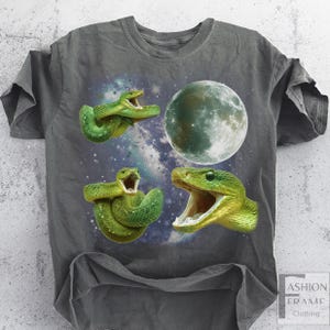May include: Dark gray t-shirt featuring a graphic design of three green snakes with open mouths, a full moon, and a starry galaxy background. The shirt has a crew neck and short sleeves.