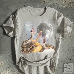 May include: A light gray t-shirt featuring a graphic of three snakes with open mouths, a full moon, and a starry background. The snakes are in shades of brown and yellow. The shirt has a crew neck and short sleeves.
