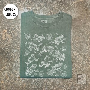 May include: Green t-shirt with a crew neck featuring a white illustration of frogs and leaves. The shirt has the words "Comfort Colors" in a white circle in the upper left corner. The shirt is folded neatly.